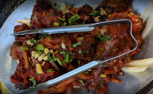 A plate of spicy meat with sauce, garnished with herbs and served with tongs on the side.