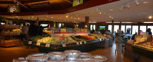 A vibrant market interior filled with fresh produce, baked goods, and shoppers browsing the aisles.