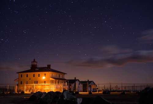 A historic lighthouse illuminated at night, surrounded by stars and a clear sky, with a fence in the background.