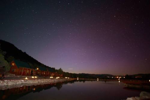 A serene night sky filled with stars reflects on a calm lake, with silhouettes of trees and buildings in the background.