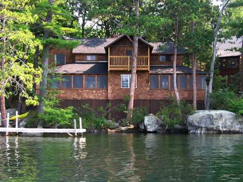 A wooden cabin by a lake, surrounded by trees, with a balcony and a dock in the foreground.