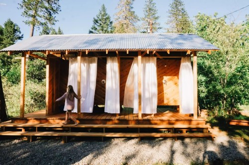 A woman walks past a wooden cabin with white curtains, surrounded by trees and gravel. Sunlight filters through.