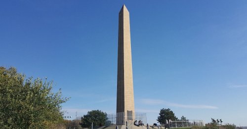 A tall stone obelisk stands against a clear blue sky, surrounded by greenery and people sitting nearby.