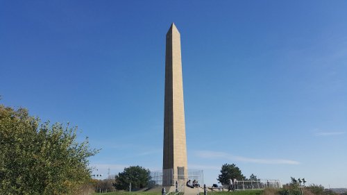 A tall, stone obelisk stands against a clear blue sky, surrounded by greenery and a few people sitting nearby.
