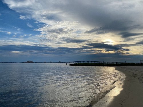 A serene beach scene at sunset, with calm water reflecting clouds and a distant pier under a cloudy sky.