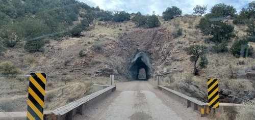 A dirt road leading into a rocky tunnel surrounded by dry hills and sparse vegetation.