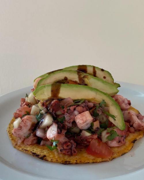 A colorful taco topped with diced octopus, avocado slices, tomatoes, and herbs on a corn tortilla.