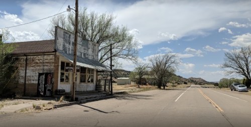 Abandoned storefront beside a deserted road, with sparse trees and mountains in the background under a cloudy sky.