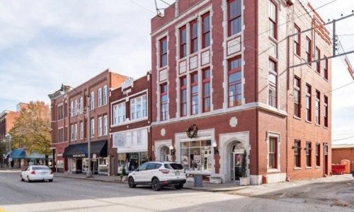 A historic brick building stands on a street lined with shops and trees, under a clear sky.