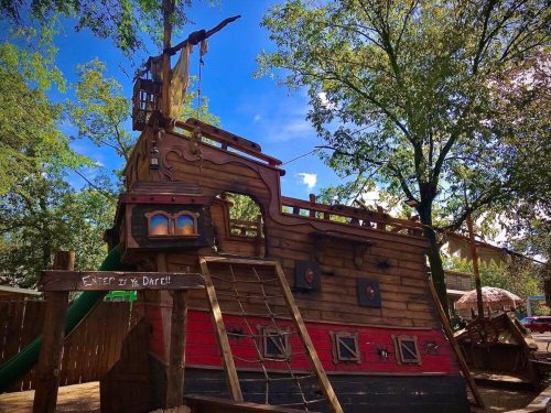A colorful wooden pirate ship playground with a slide, surrounded by trees and a blue sky.