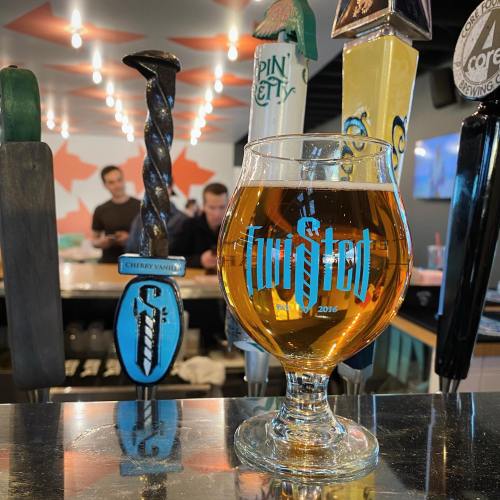 A glass of amber beer sits on a bar, with colorful tap handles in the background and patrons chatting.