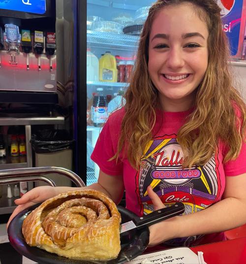 A smiling young woman holds a large cinnamon roll on a plate in a diner setting.