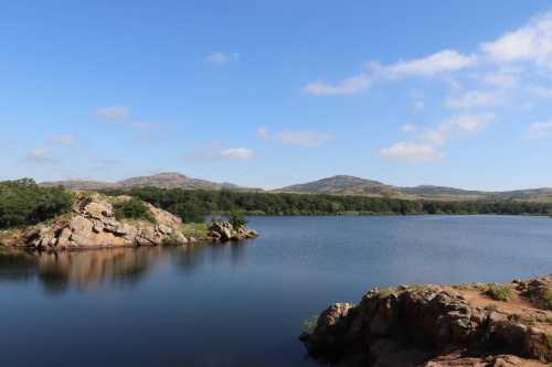 A serene lake surrounded by rocky shores and lush greenery, with mountains in the background under a clear blue sky.