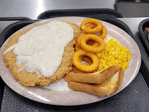 A plate with fried chicken smothered in gravy, onion rings, corn, and two slices of toasted bread.