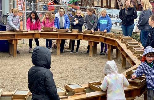 Children gather around a wooden structure, engaged in an activity, with adults supervising in the background.