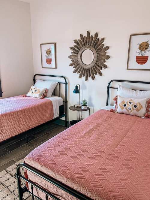 A cozy bedroom with two metal frame beds, coral bedding, decorative pillows, and a round mirror on the wall.