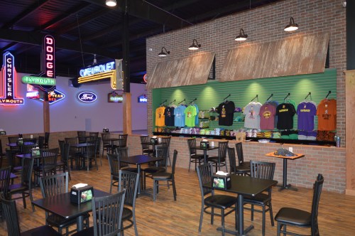 Interior of a restaurant featuring tables, neon signs, and a display of colorful t-shirts on a green wall.