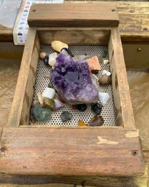 A wooden box filled with various colorful gemstones and crystals, including a large purple amethyst.