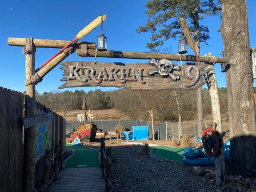 Sign for "Kraken" with a skull and crossbones, set in a playful outdoor mini-golf area surrounded by trees.
