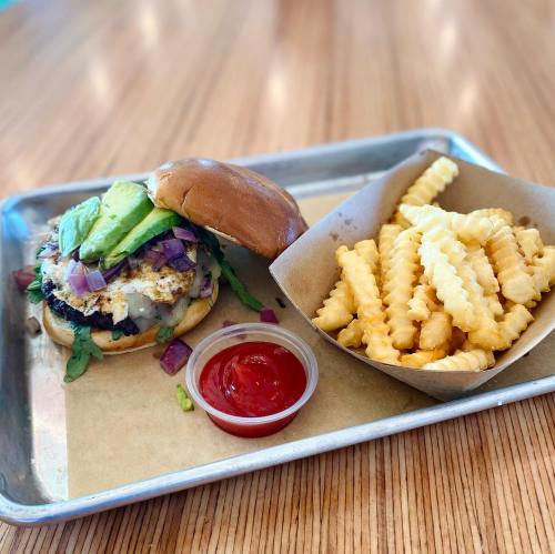 A burger with avocado, onions, and greens on a bun, served with crinkle-cut fries and ketchup on a metal tray.