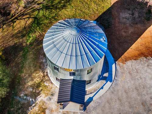 Aerial view of a round metal silo with a conical roof, surrounded by grass and gravel.