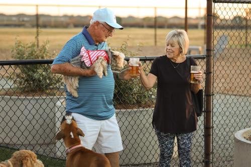 An older man and woman toast with drinks while holding small dogs at a park, with a dog nearby.