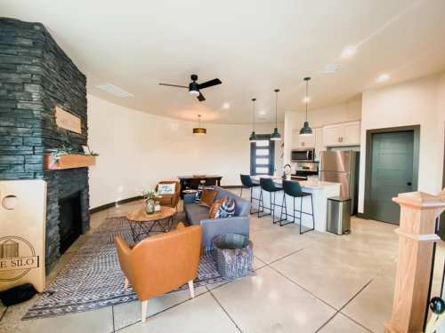 Modern living room with a stone fireplace, dining area, and kitchen, featuring stylish furniture and bright lighting.