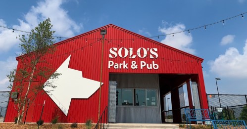 Red metal building with "SOLO's Park & Pub" sign and a large white arrow, under a blue sky with scattered clouds.
