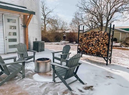 A cozy outdoor seating area with four chairs around a fire pit, snow on the ground, and a wood stack nearby.