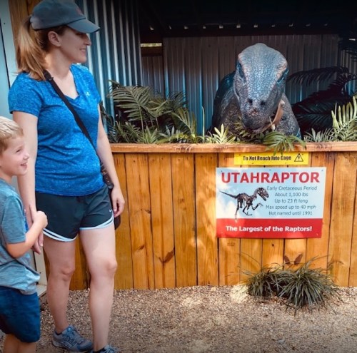 A woman and a boy stand near a large dinosaur exhibit labeled "Utahraptor," with greenery in the background.