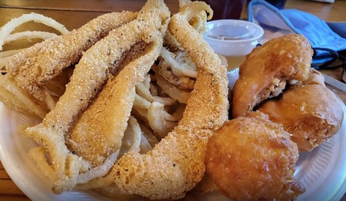 A plate of fried fish fillets, onion rings, a hushpuppy, and a fried pastry, served with dipping sauce.