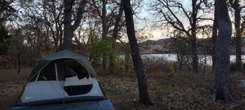 A camping tent set up among trees, with a serene lake and autumn foliage in the background at dusk.