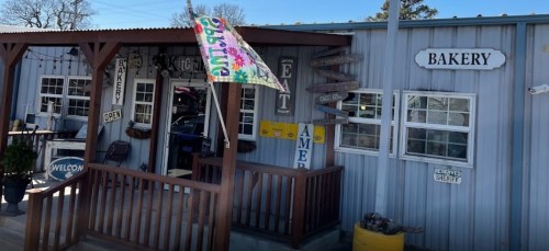 A charming bakery with a welcoming porch, colorful flag, and directional signs, set in a rustic metal building.