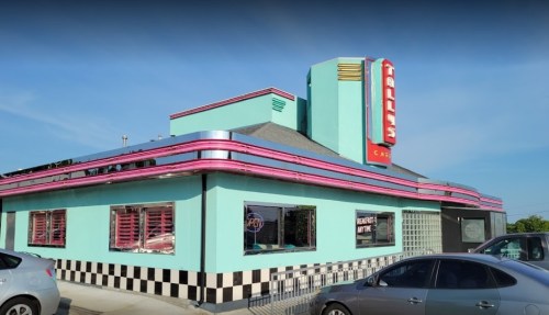 A retro-style diner with a turquoise exterior, pink accents, and a neon sign, surrounded by parked cars.