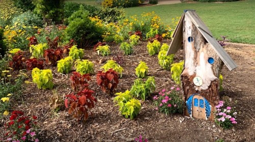 A whimsical wooden fairy house surrounded by colorful plants and flowers in a garden setting.