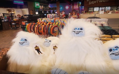 Three fluffy yeti plush toys are displayed in a colorful gift shop filled with various items in the background.