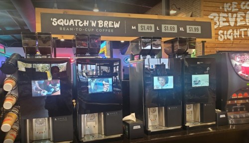 Self-service coffee machines labeled 'Squatch'n Brew' with pricing displayed above. Screens show various images.