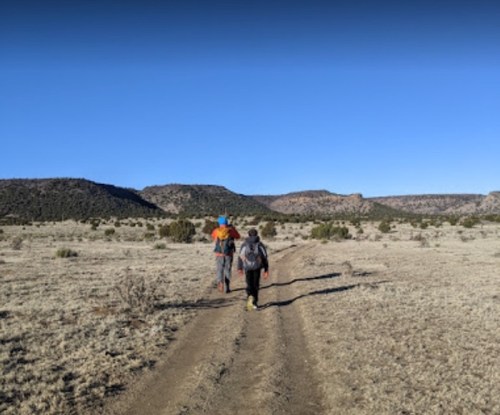 Two hikers walking on a dirt path through a dry, open landscape with distant hills under a clear blue sky.