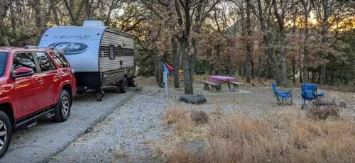 A red SUV parked next to a camper in a wooded area, with a picnic table and two blue chairs nearby.