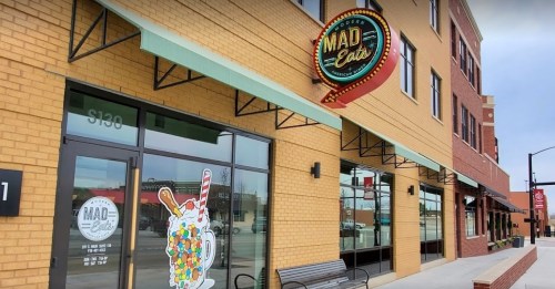 Exterior of Mad Eats restaurant, featuring a colorful sign and large windows, with a bench outside.