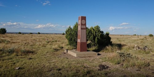 A tall, red monument stands on a grassy plain under a blue sky, surrounded by sparse vegetation and distant hills.