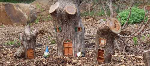 Whimsical tree stump houses with tiny doors and windows, surrounded by natural foliage and decorative garden gnomes.