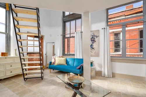 Bright, modern living space featuring a blue sofa, glass table, and a stylish staircase under large windows.