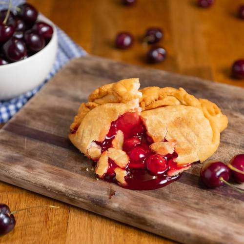 A cherry-filled pastry with a flaky crust, partially broken open, surrounded by fresh cherries on a wooden surface.