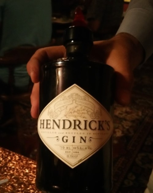 A hand holding a bottle of Hendrick's Gin, featuring a distinctive black label and red cap.