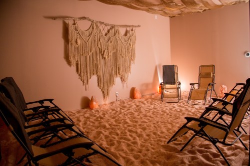 A serene room with a sandy floor, two chairs, and a macramé wall hanging, creating a calming atmosphere.