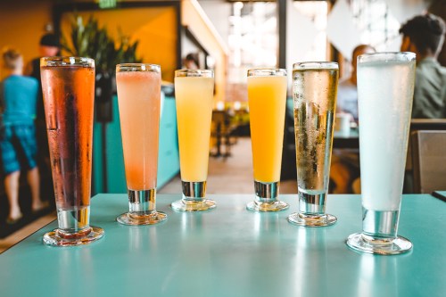 A row of six colorful drinks in tall glasses on a table, with a bright, modern café setting in the background.