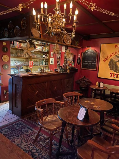 A cozy bar interior featuring wooden furniture, a chandelier, and a well-stocked counter with decorative wall elements.