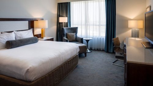 A modern hotel room featuring a king-sized bed, armchair, desk, and large window with sheer curtains.