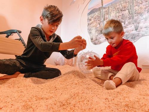 Two children play together on a soft, sandy surface, focusing on a clear, round object between them.
